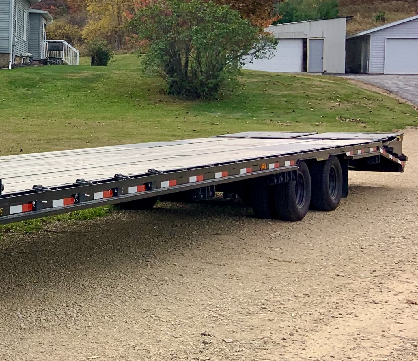 PLACEHOLDER: the timber deck, dovetail and tandem axles of a gooseneck flatbed trailer standing on a gravel drive.